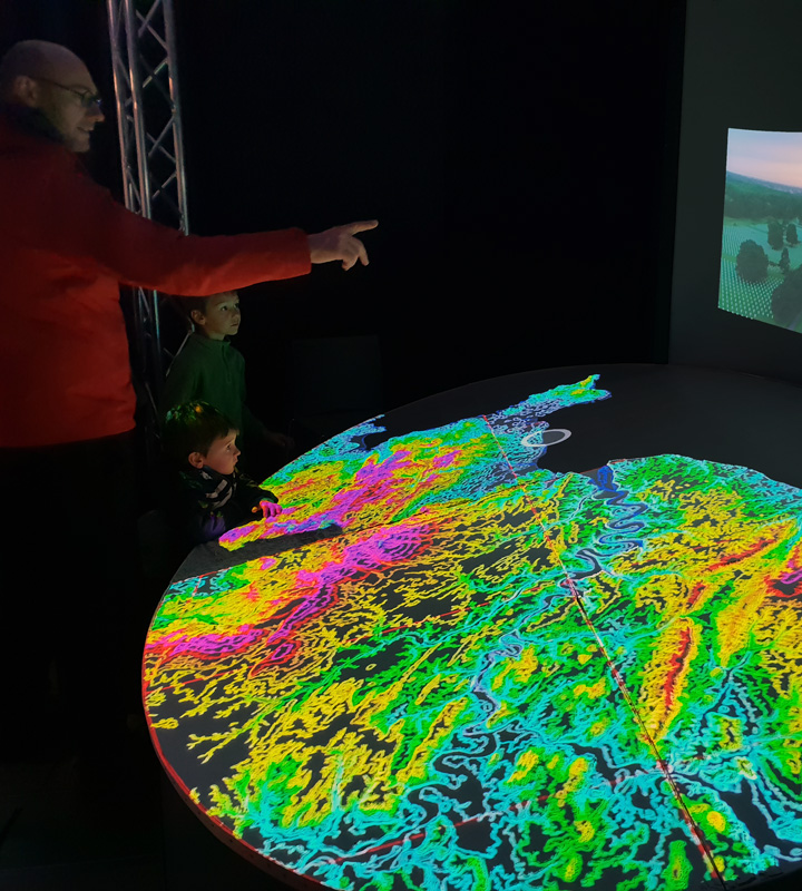 Lors d'un événement, un homme et ses deux jeunes fils discutent et pointent du doigt la maquette interactive NormanDisplay.